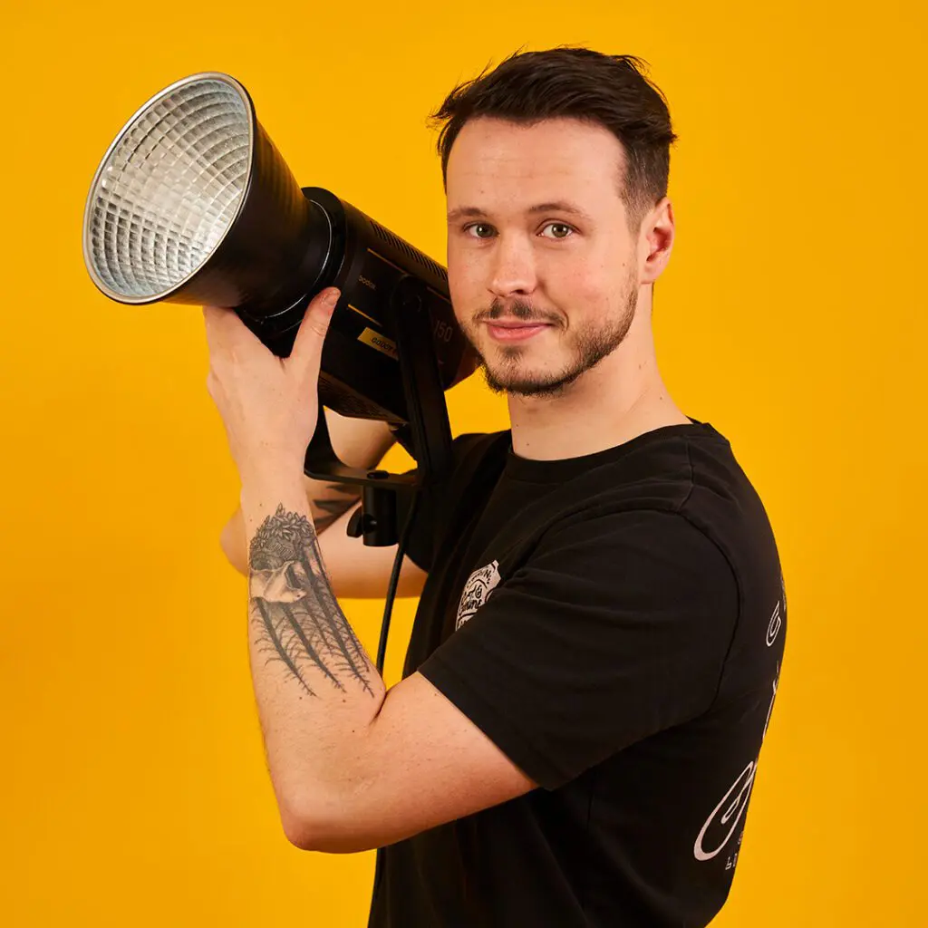 Chris - man with short dark hair and beard wearing black tshirt with arm tattoo holding a spotlight