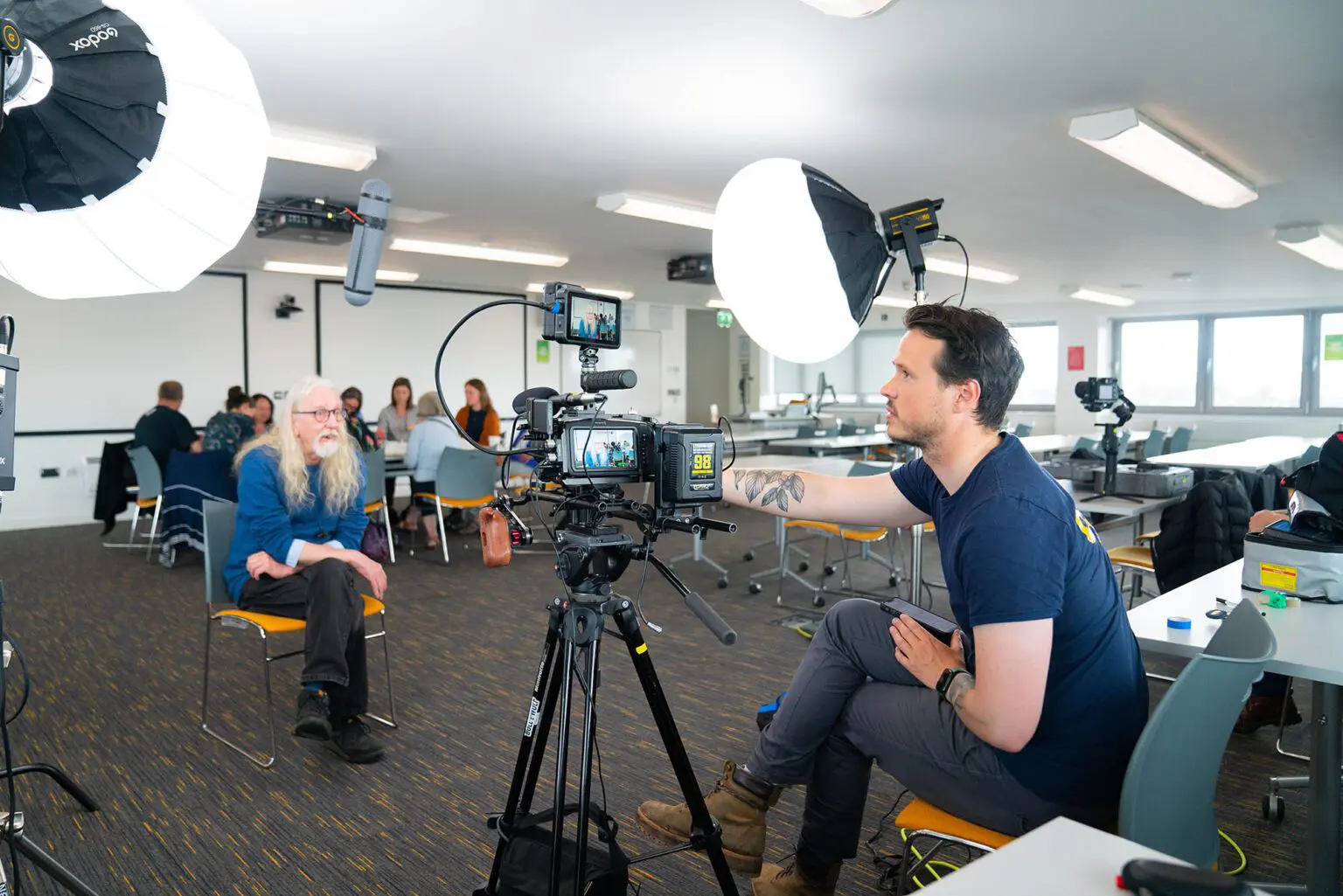 Behind the camera angle, showing camera screens and Chris using the camera. Man sat on chair in front of lense.