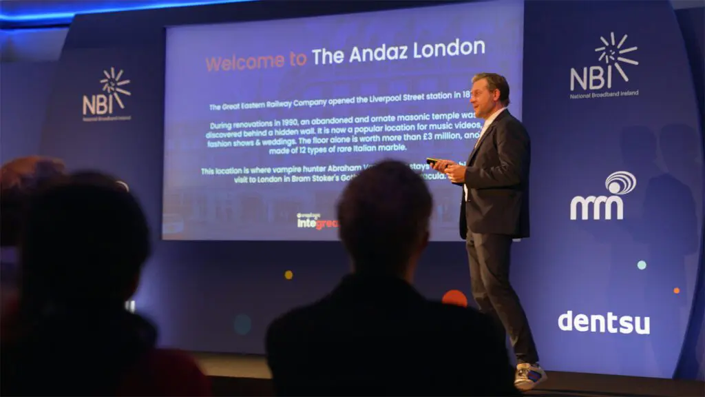 Man in suit stood on stage at a presenting slides