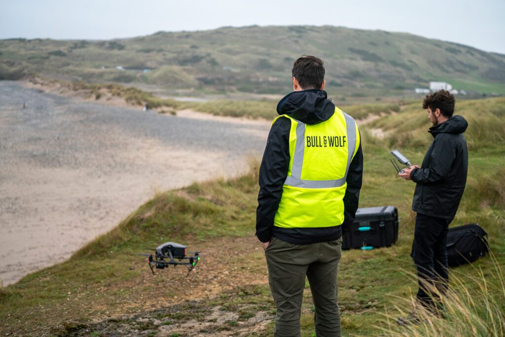 Drone filming shoot. Two men one in high vis and one with controls looking at drone in sanddunes