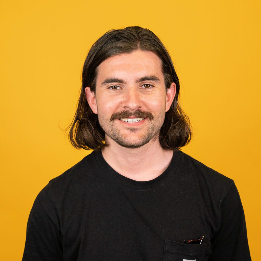 Man headshot in black tshirt with sunglasses in pocket with shoulder length brown hair and moustache